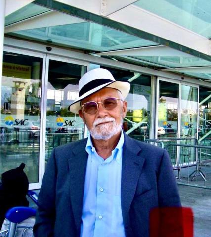 This photo shows a distinguished gentleman wearing a white fedora and sunglasses, exuding a relaxed and confident demeanor as he stands outside what appears to be an airport or transportation hub. The background includes glass doors and signs, adding to the travel setting.